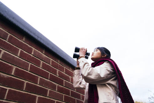 Frau mit Fernglas in der Nähe einer Backsteinmauer stehend - ASGF02062