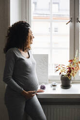 Pregnant woman with hand on stomach looking through window at home - MASF28049