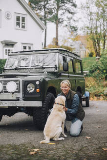 Lächelnde reife Frau streichelt Hund an einem Geländewagen in der Einfahrt - MASF28109