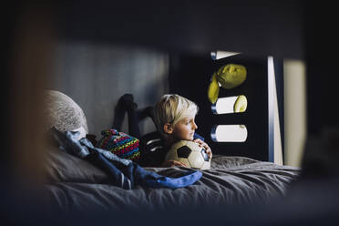 Boy with soccer ball looking away while lying on bed - MASF28165