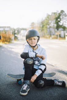 Boy sitting on skateboard at road - MASF28221