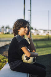 Female athlete with soccer ball sitting on retaining wall - MASF28231