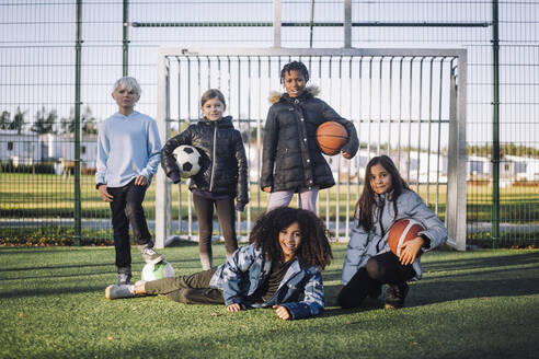 Portrait of girls and boy with sports balls on soccer field - MASF28233