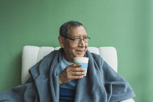 Smiling senior man with disposable coffee cup in front of green wall - DSIF00587