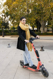 Smiling teenage girl riding electric push scooter on footpath - JRVF02362