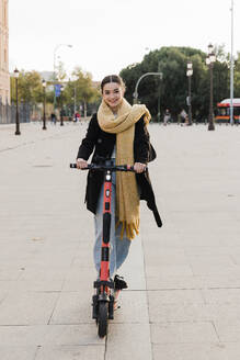 Happy teenager riding electric push scooter on footpath - JRVF02363