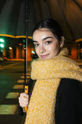 Lächelndes Teenager-Mädchen mit Schal und Regenschirm auf der Straße bei Nacht - JRVF02371