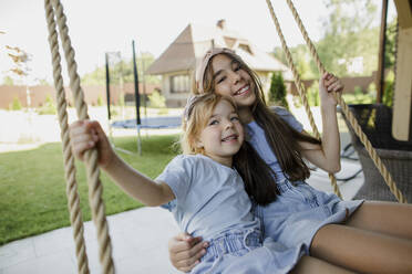 Happy sisters sitting on swing at patio - LLUF00443