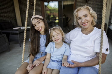 Smiling grandmother with granddaughters sitting on rope swing - LLUF00445