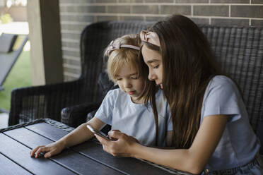 Girl sharing smart phone with sister at patio - LLUF00451