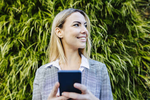 Junge blonde berufstätige Frau mit Smartphone vor Grünpflanzen - XLGF02504