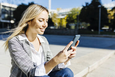 Smiling blond working woman using smart phone on sunny day - XLGF02507