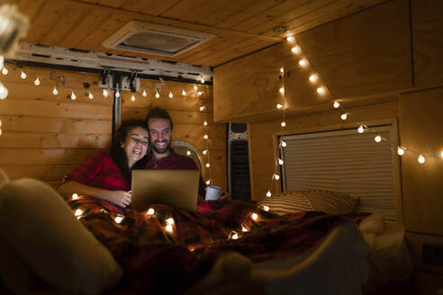 Glückliches Paar, das in einem beleuchteten Wohnmobil einen Film auf dem Laptop ansieht - EGHF00297