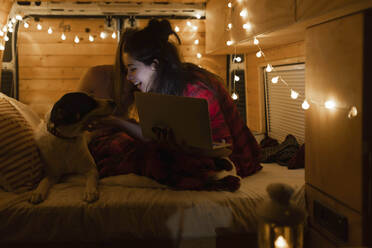 Happy woman with laptop looking at dog in illuminated van - EGHF00306