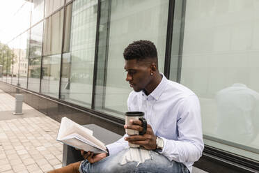 Junger Mann mit wiederverwendbarem Kaffeebecher, der vor einem Gebäude ein Buch liest - VYF00874