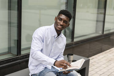 Smiling young man with book sitting on bench - VYF00877