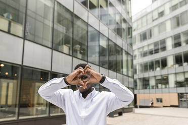 Young man making heart shape in front of building - VYF00885