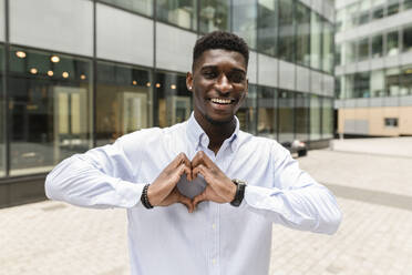 Smiling man gesturing heart shape on footpath - VYF00886