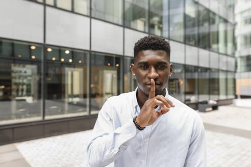 Junger Mann mit Finger auf den Lippen vor einem Glasgebäude - VYF00888