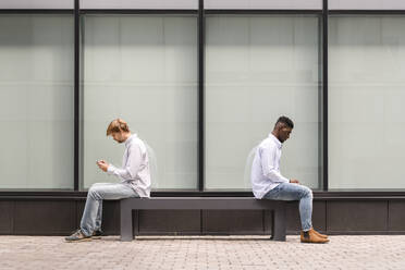 Freunde sitzen Rücken an Rücken auf einer Bank und benutzen ein Smartphone - VYF00907