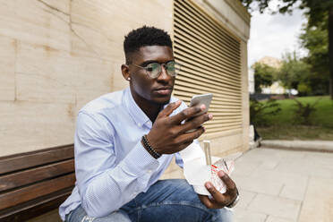 Junger Mann mit Brotdose und Smartphone auf einer Bank - VYF00930