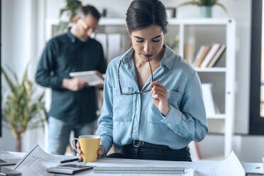 Businesswoman having coffee while working on floor plan at workplace - JSRF01780