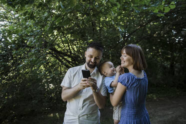 Smiling woman carrying son and looking at man showing smart phone - LLUF00454