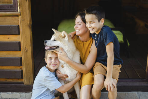 Glückliche Familie umarmt ihren Akita-Hund, der vor dem Haus sitzt - SEAF00325