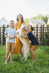 Cheerful family with their Akita dog standing on grass in front of fence - SEAF00337