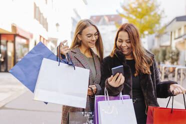 Glückliche Frauen mit Einkaufstüten, die auf dem Fußweg ihr Smartphone teilen - DAWF02380