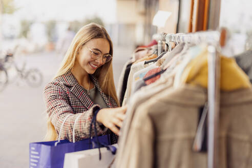 Smiling woman shopping clothes arranged on rack outside store - DAWF02385