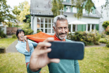 Cheerful woman pulling scarf of man taking selfie on mobile phone - JOSEF06402