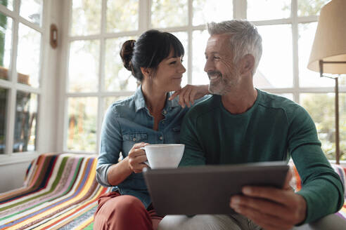 Lächelnder Mann mit Tablet-PC neben einer Frau mit Kaffeetasse auf dem Sofa - JOSEF06413