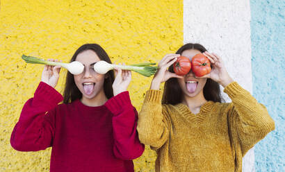 Verspielte Schwestern bedecken ihre Augen mit Gemüse vor einer bunten Wand - JCCMF04990