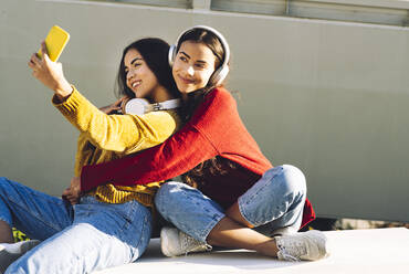 Glückliche Schwestern mit Kopfhörern nehmen Selfie zusammen auf Bank - JCCMF05001