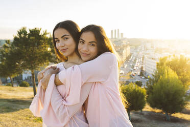 Smiling sisters embracing each other in park at sunset - JCCMF05014