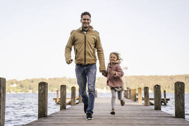 Smiling father holding hand of daughter and running on jetty - DIGF17374