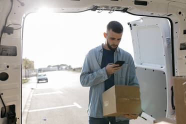 Zusteller, der ein Mobiltelefon benutzt und eine Box an der Tür des Lieferwagens hält - JRFF05202