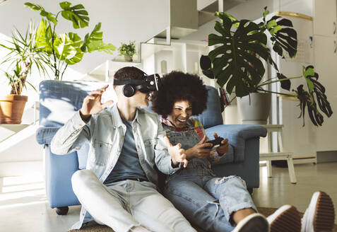 Junger Mann mit VR-Brille gestikuliert bei fröhlicher Freundin, die zu Hause ein Videospiel spielt - JCCMF05032