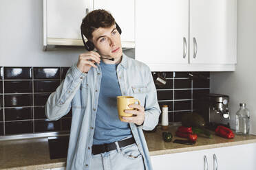 Junger Mann mit Headset und Kaffeetasse auf dem Küchentisch zu Hause - JCCMF05045