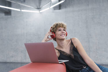 Smiling businesswoman listening music wih headphones by laptop in office - PNAF02727