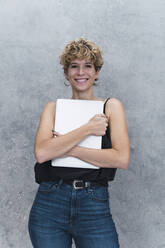 Lächelnde junge Geschäftsfrau mit Laptop vor einer grauen Wand - PNAF02729