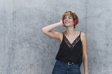 Lächelnde Frau hört Musik über Kopfhörer vor einer grauen Wand - PNAF02731