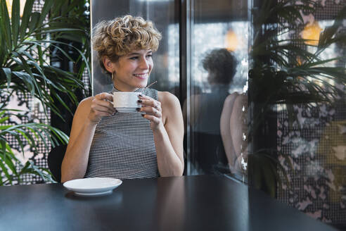 Lächelnde junge Frau mit Kaffeetasse am Tisch in einem Café sitzend - PNAF02737