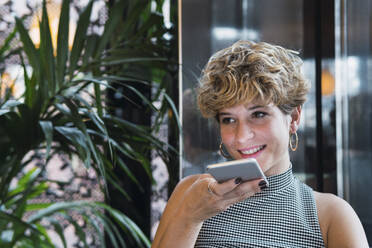 Lächelnde Geschäftsfrau, die in einem Café eine Voicemail auf ihrem Smartphone verschickt - PNAF02744