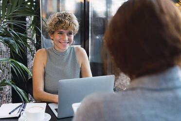 Junge Geschäftsfrau mit Laptop, die einen Kollegen in einem Café anlächelt - PNAF02751