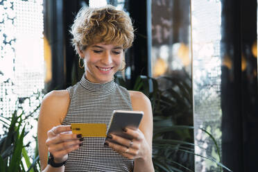 Lächelnde junge Frau benutzt Kreditkarte und Smartphone zum Online-Shopping in einem Café - PNAF02756