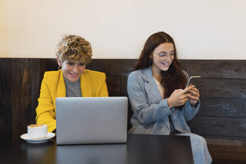 Lächelnde Geschäftsfrau, die in einem Café mit einem Kollegen am Laptop arbeitet und ein Smartphone benutzt - PNAF02766