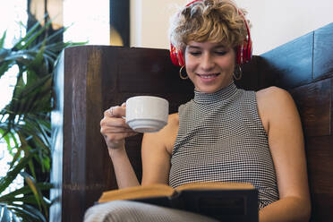 Lächelnde Frau mit Kopfhörern, die in einem Café Kaffee trinkt und ein Buch liest - PNAF02767