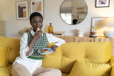 Thoughtful woman eating fruits on sofa at home - JCZF00882